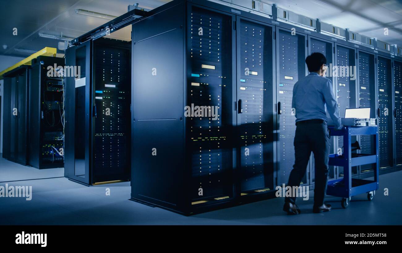 Nel moderno data center: Tecnico IT che esegue la manutenzione e la diagnostica. Spinge il carrello accanto ai rack server. Aggiornamento pianificato delle apparecchiature di sistema e. Foto Stock
