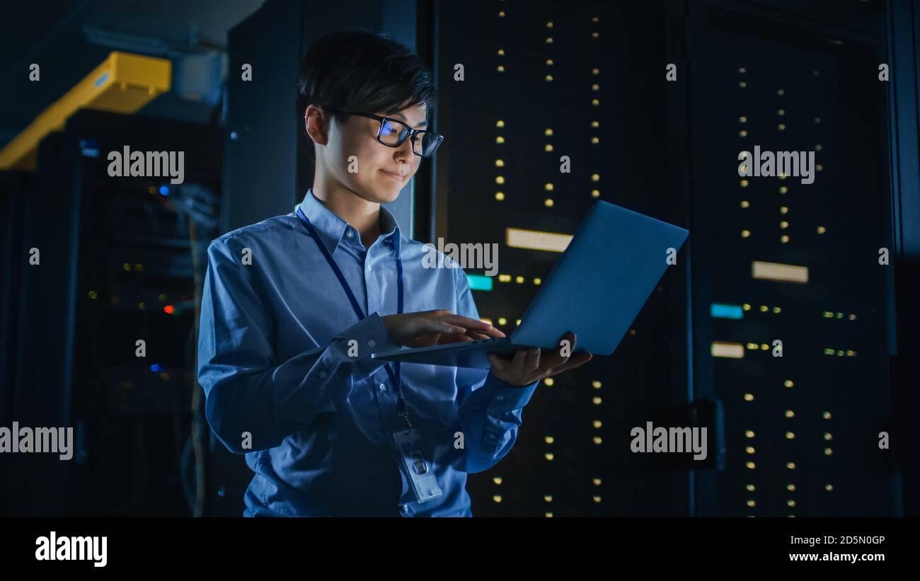 Nei data center bui: Lo specialista IT maschile si trova accanto alla riga dei rack server operativi e utilizza il notebook per la manutenzione. Concetto per il cloud computing Foto Stock