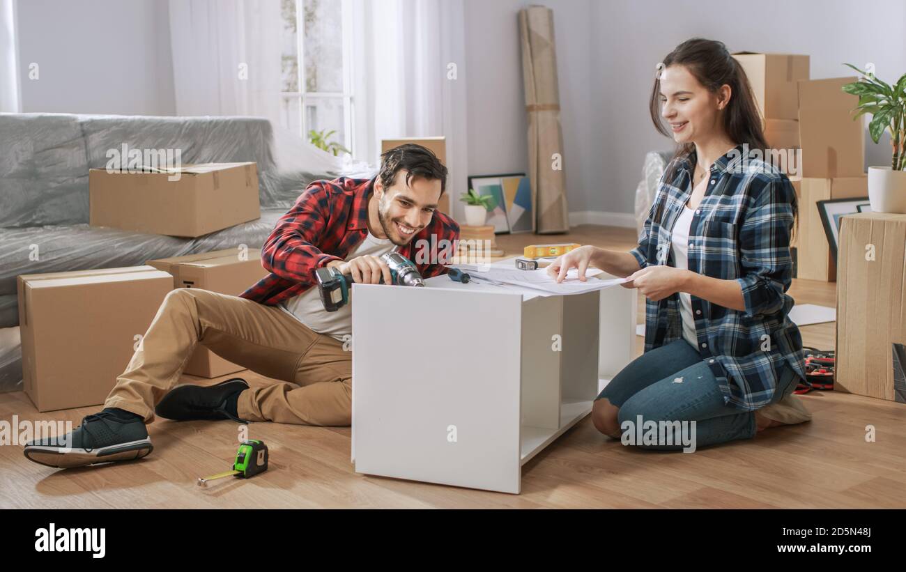 Felice coppia che monta mobili come squadra, ragazza legge istruzioni e ragazzo tensiona vite con un trapano. Spostandosi in nuovo appartamento, insieme di coppia Foto Stock