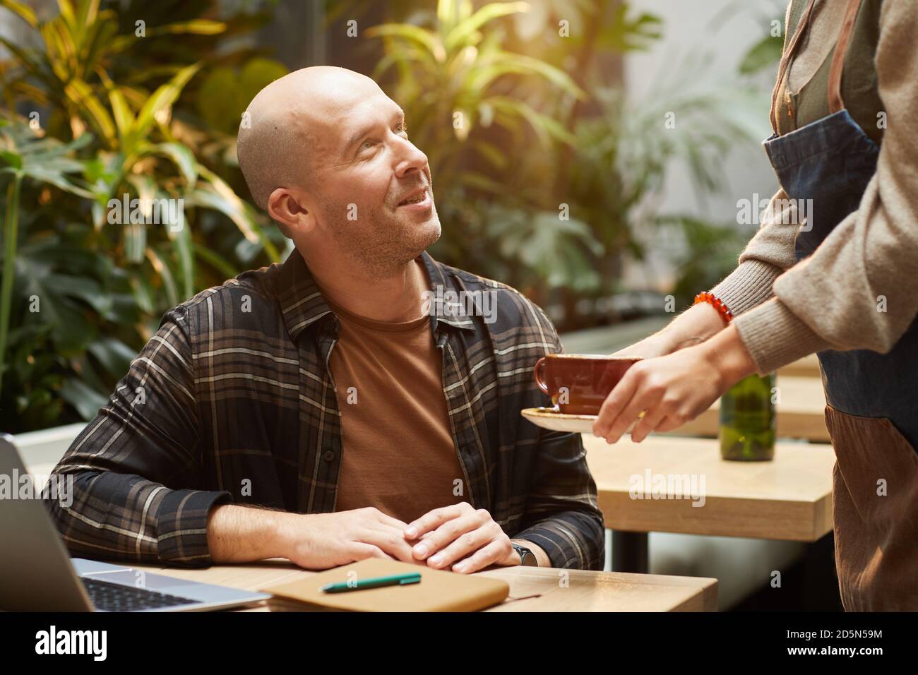 Uomo maturo seduto al tavolo con il computer portatile e che parla al cameriere mentre serve il caffè per lui in caffetteria Foto Stock