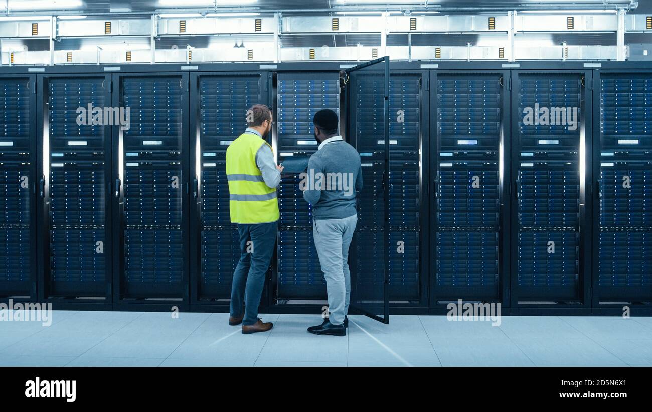 Amministratore IT con un computer portatile in High Visibility Vest E Black Male Engineer stanno parlando in data center mentre Davanti a un rack server Foto Stock
