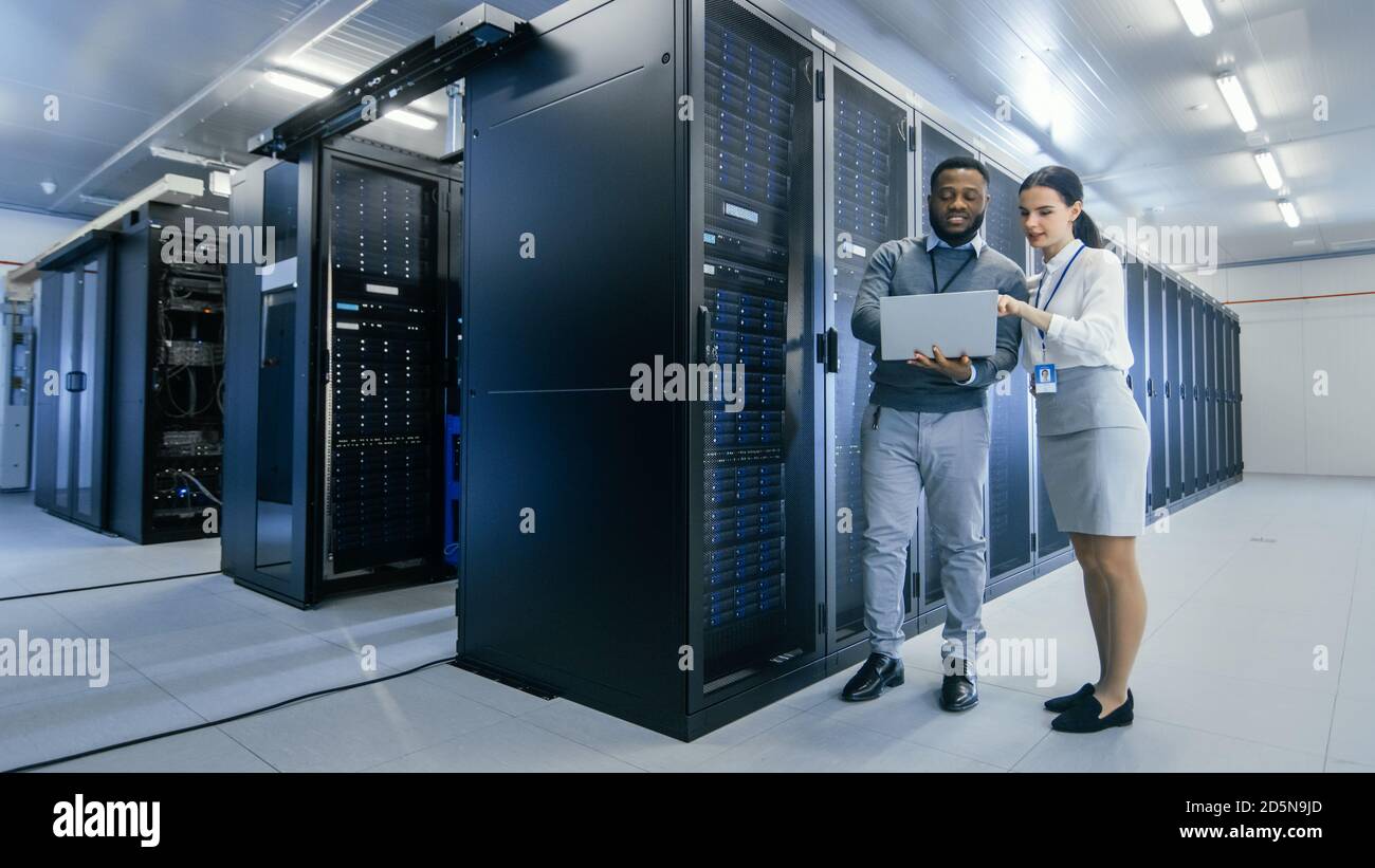 Black IT Technician con un computer portatile offre un tour a un giovane internista. Parlano nel data center mentre si cammina accanto ai rack server. In esecuzione Foto Stock