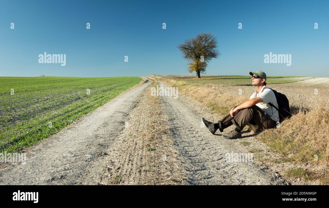 Un uomo seduto accanto alla strada, campi e un albero solitario Foto Stock