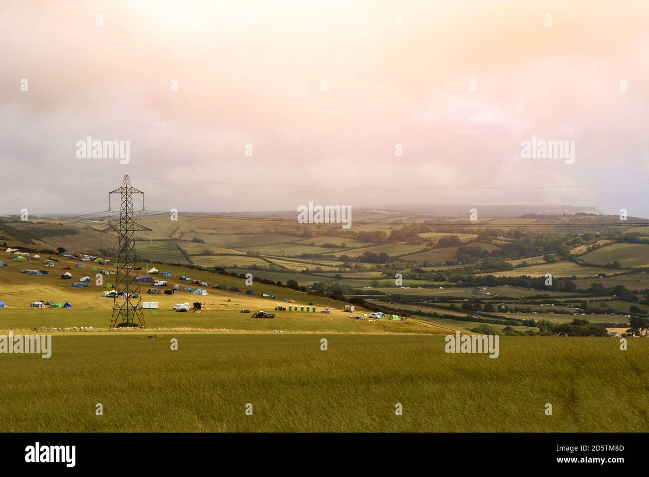 Una vista sulla campagna del Dorset con un campeggio di tende in lontananza e una torre elettrica a reticolo. Foto Stock