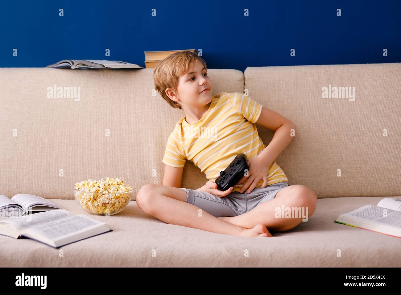 Il ragazzo di scuola biondo gioca i video giochi, tiene un gamepad, mangia il popcorn invece di imparare le lezioni. Foto Stock