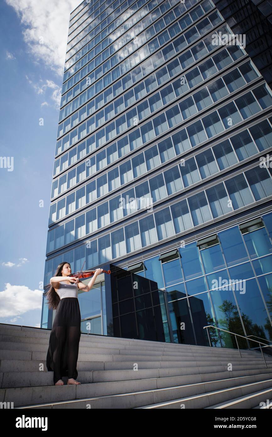 Giovani asiatici femmina suona il violino su scale davanti a un edificio alto e moderno Foto Stock