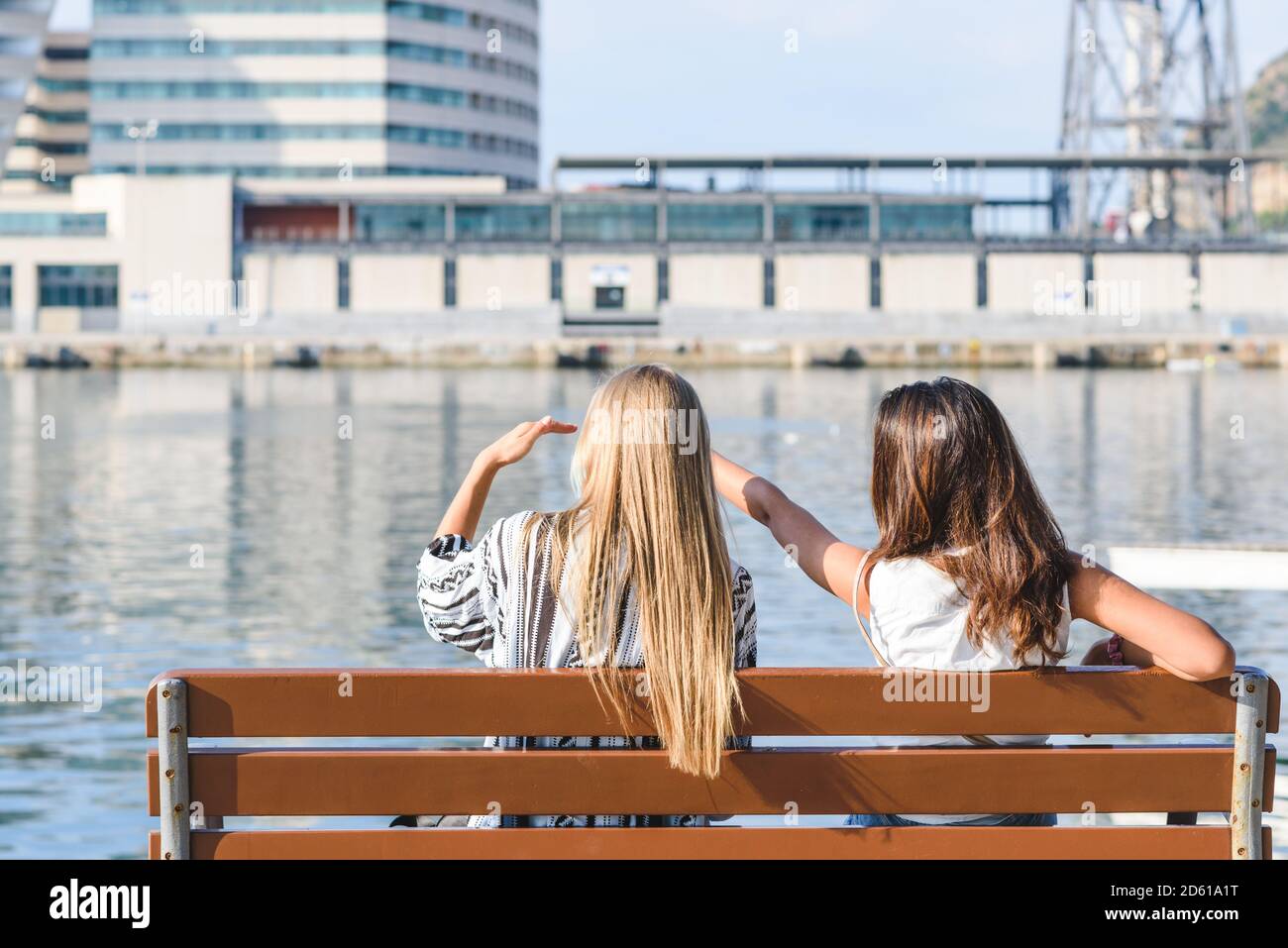 vista posteriore di due amici femminili seduti su una panchina e indicando qualcosa che cattura il loro interesse in un porto area Foto Stock