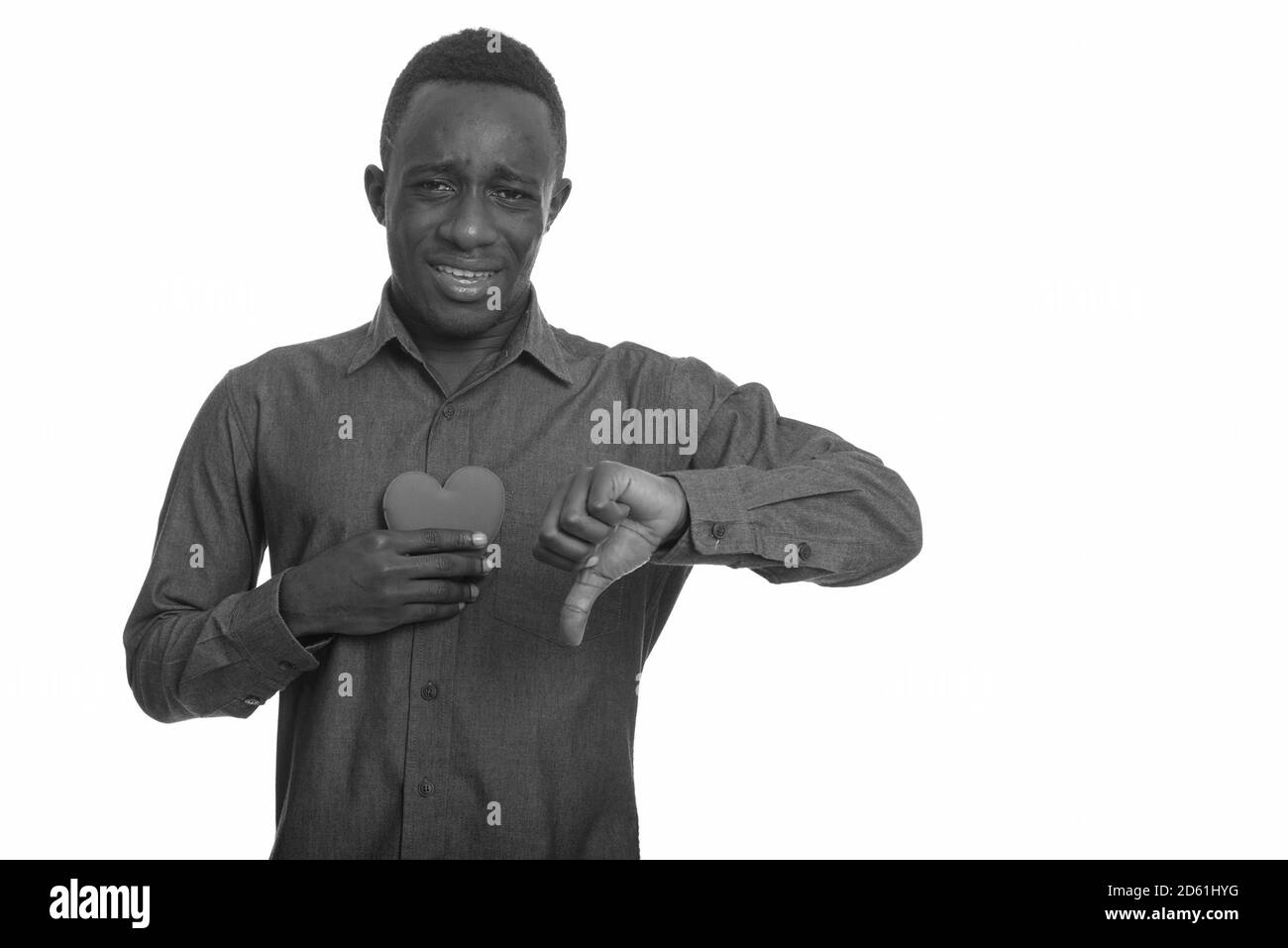 Giovane uomo africano triste che tiene il cuore sul petto e dà pollice giù Foto Stock