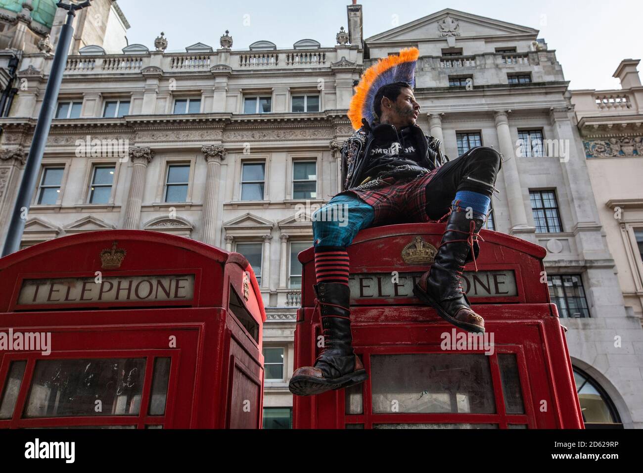 Punk rocker con capelli mohican si siede rilassante sulla parte superiore delle scatole rosse del telefono nel centro di Londra, Inghilterra, Regno Unito Foto Stock