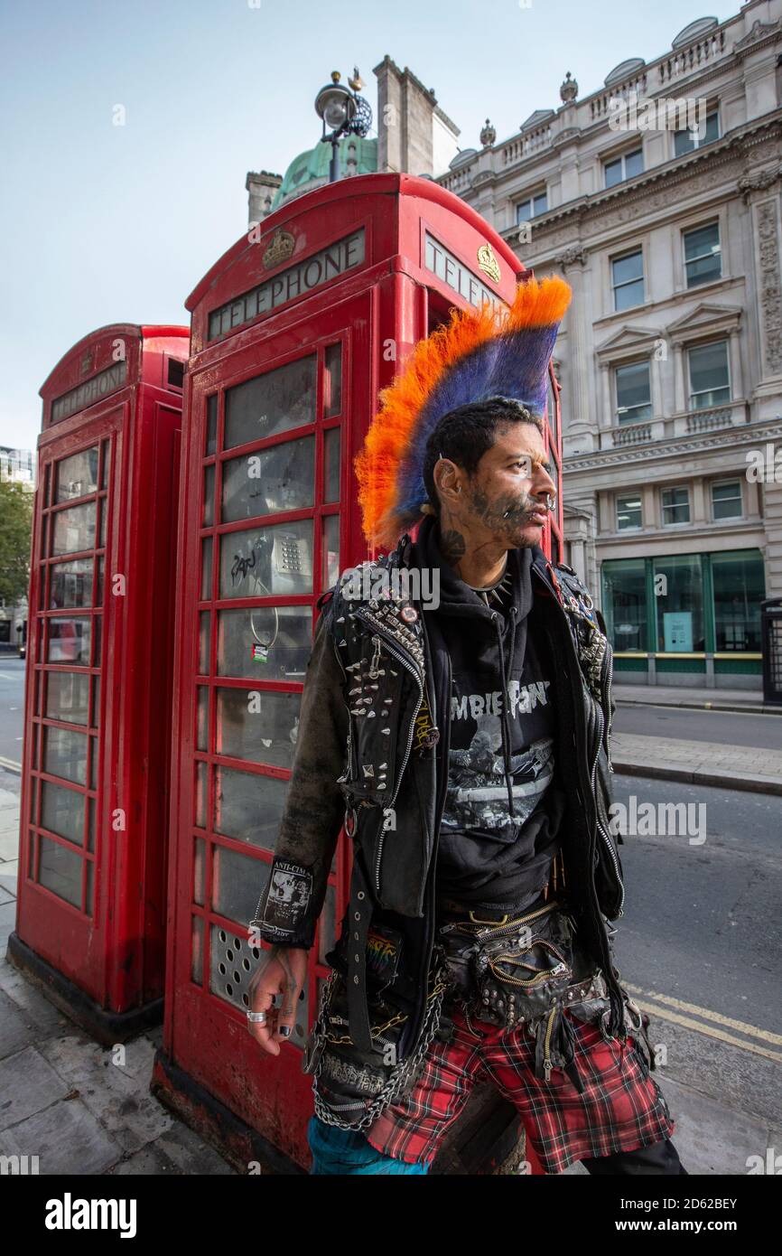 Punk rocker con capelli mohican si siede rilassante sulla parte superiore delle scatole rosse del telefono nel centro di Londra, Inghilterra, Regno Unito Foto Stock