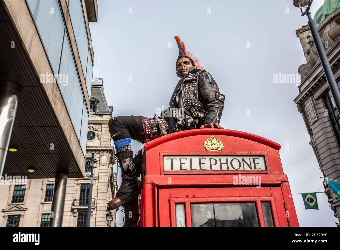 Punk rocker con capelli mohican si siede rilassante sulla parte superiore delle scatole rosse del telefono nel centro di Londra, Inghilterra, Regno Unito Foto Stock