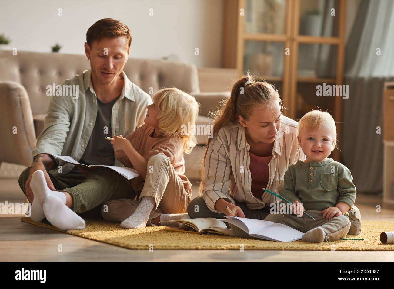 Due genitori seduti sul pavimento e leggendo un libro ai loro figlioli nel soggiorno di casa Foto Stock
