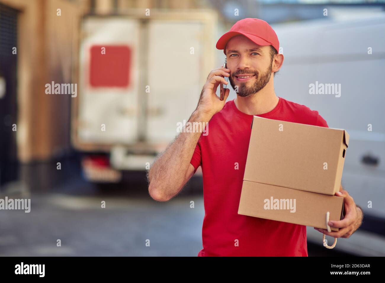 Giovane allegro Courier maschio caucasico con scatola di carta sorridente a macchina fotografica mentre parla al telefono nel deposito cortile. Concetto di consegna degli alimenti Foto Stock