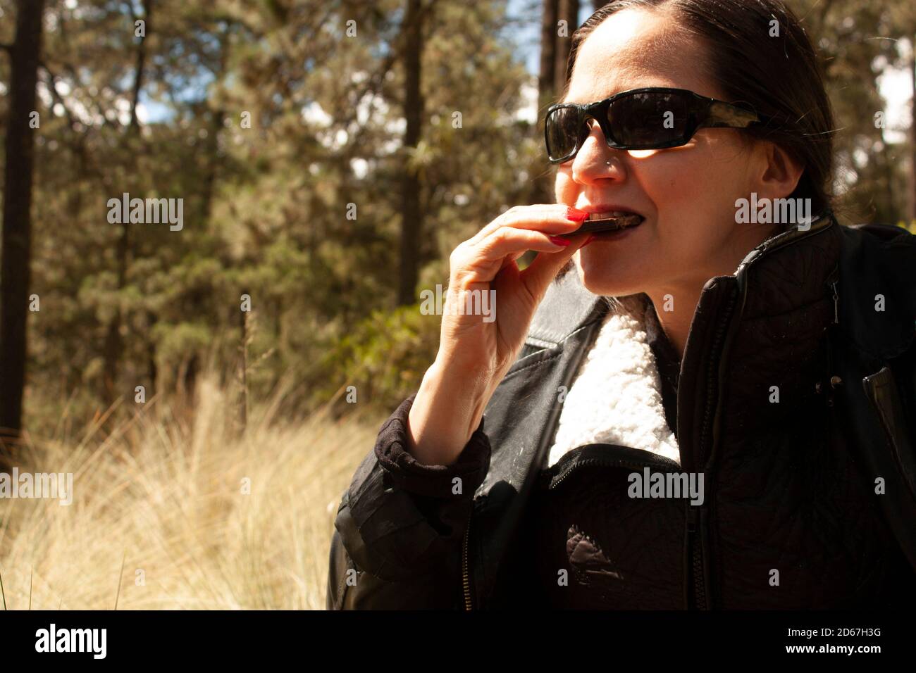 Donna che indossa occhiali da sole mangiando cioccolato che lo tiene con la mano che mostra Unghie dipinte di rosso sotto la luce del sole nello stile di vita della foresta in Messico Foto Stock