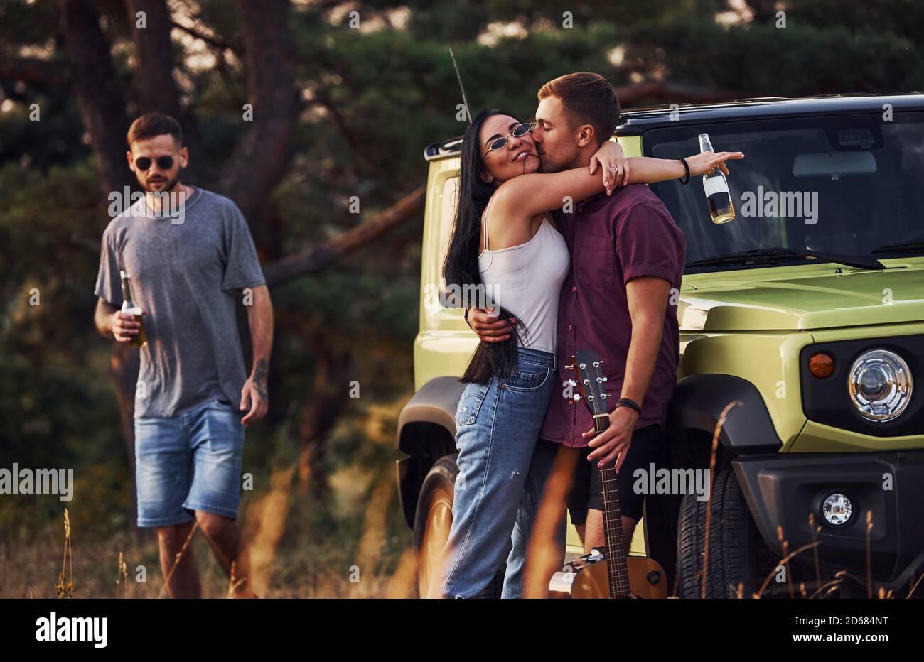 Ragazzo bacia la sua ragazza. Gruppo di amici allegri hanno fine settimana piacevole in giornata di sole vicino alla loro auto verde all'aperto Foto Stock