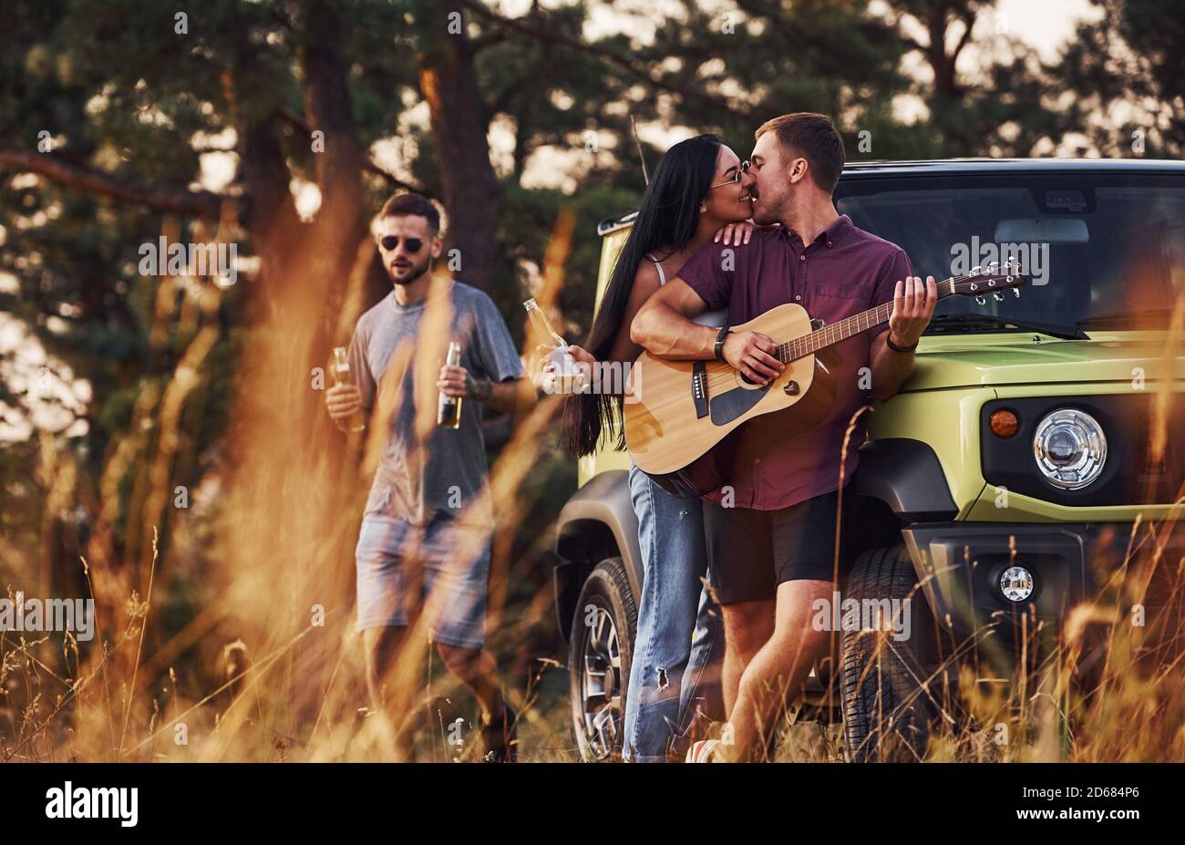 Suonando con chitarra acustica e cantando. Gruppo di amici allegri hanno fine settimana piacevole in giornata di sole vicino alla loro auto verde all'aperto Foto Stock