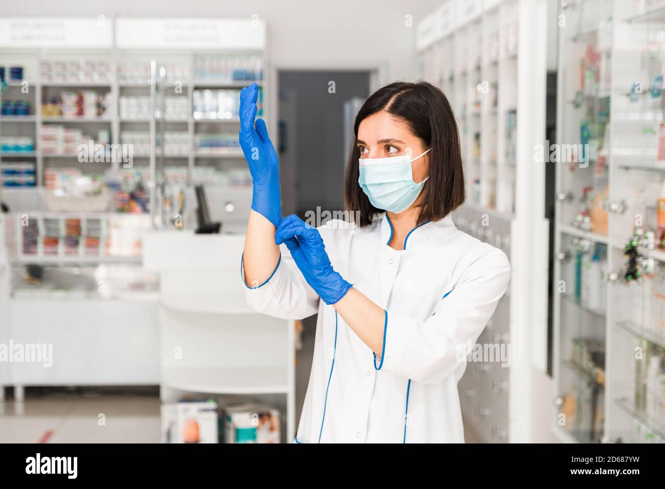 attraente grave farmacista femminile in farmacia con maschera sul viso indossare guanti blu per lavorare Foto Stock