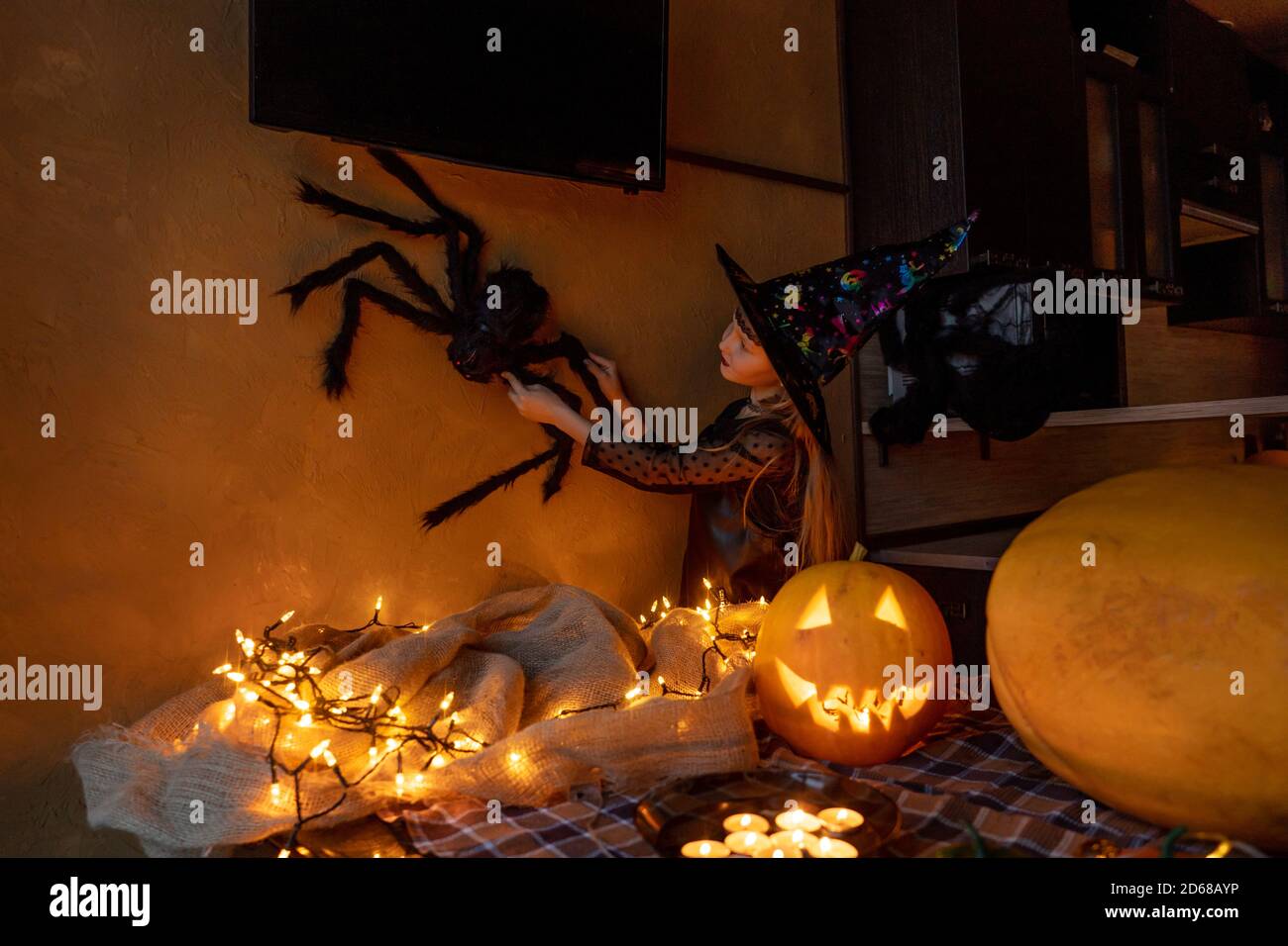 Bambina che si prepara a celebrare Halloween in costume strega. Grande ragno in mano sulla parete come arredamento. Zucca illuminata e ghirlanda sul tavolo. Foto Stock