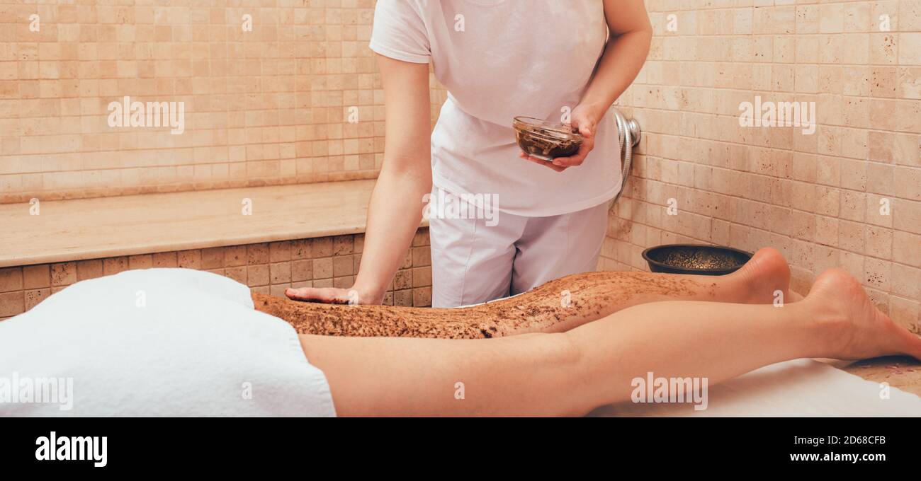 Il massaggiatore applica uno scrub di fango alle gambe di una donna. La procedura di peeling per una donna renderà le gambe lisce. Primo piano Foto Stock