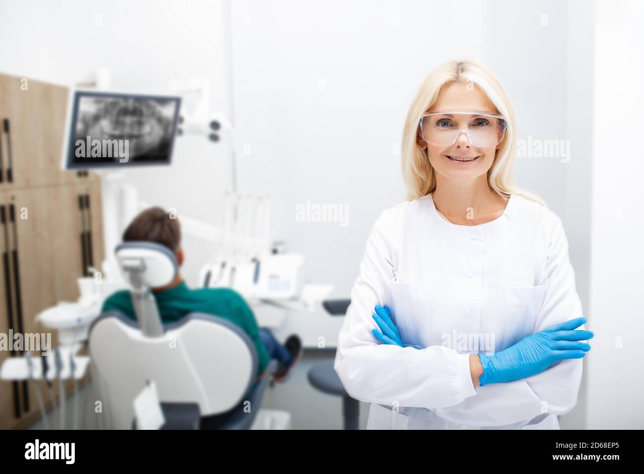 Ritratto di una donna dentista sorridente con mani incrociate, ambulatorio di odontoiatria di fondo con un paziente. Trattamento dei denti clinica moderna Foto Stock
