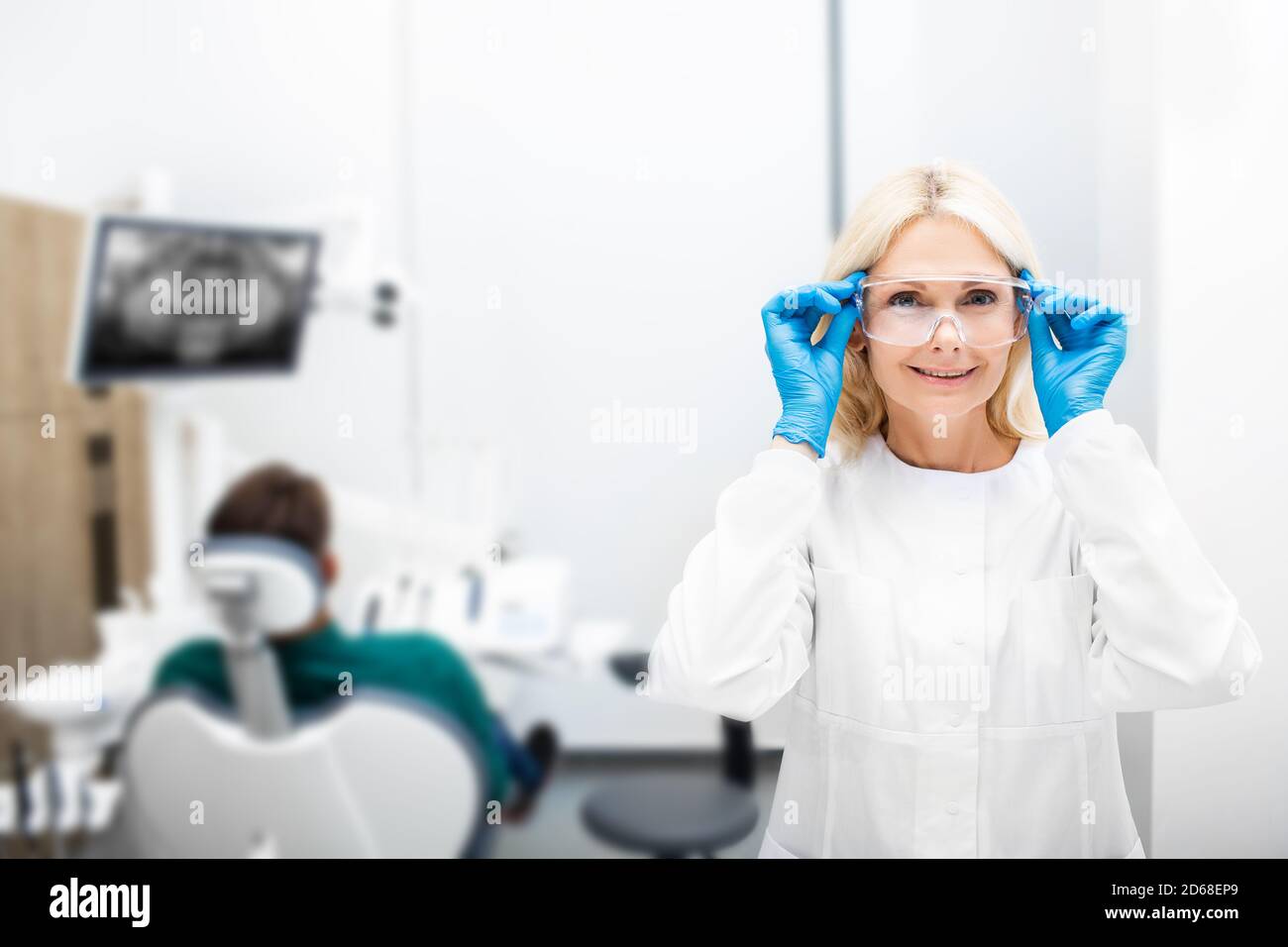 Ritratto di una donna dentista sorridente e guardando la macchina fotografica, studio di odontoiatria con un paziente. Occhiali di protezione regolabili dal dentista Foto Stock