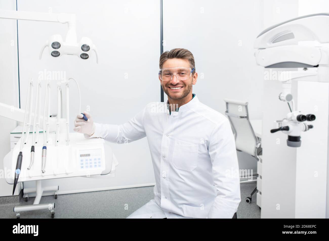 Dentista maschile sicuro seduto in una clinica moderna vicino alla sua sedia dentale. Igienista dentale che indossa occhiali protettivi sorridendo alla fotocamera Foto Stock