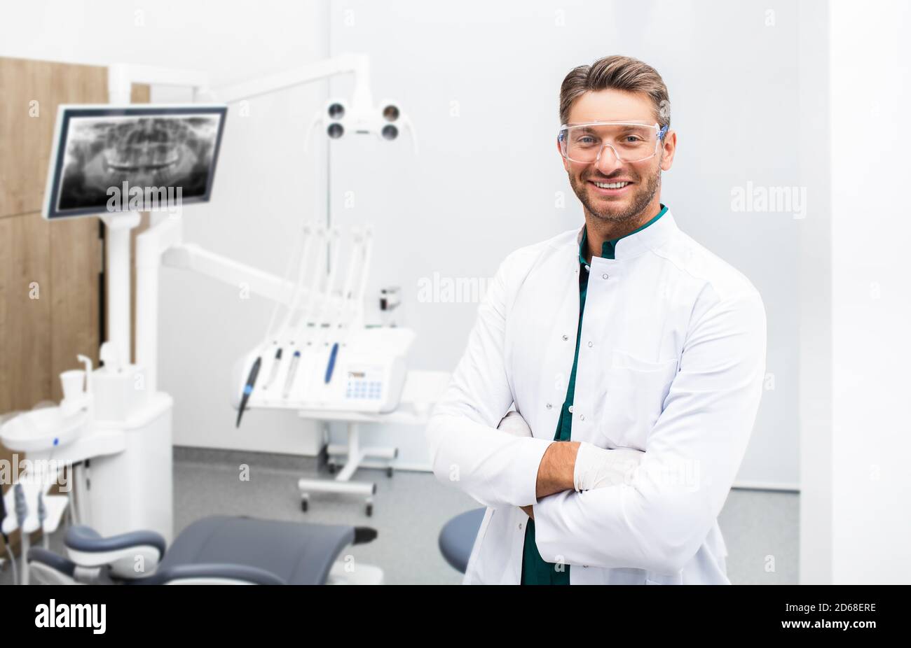 Dentista maschile sicuro in piedi nella sua clinica moderna. Igienista dentale che indossa occhiali protettivi sorridendo alla fotocamera Foto Stock