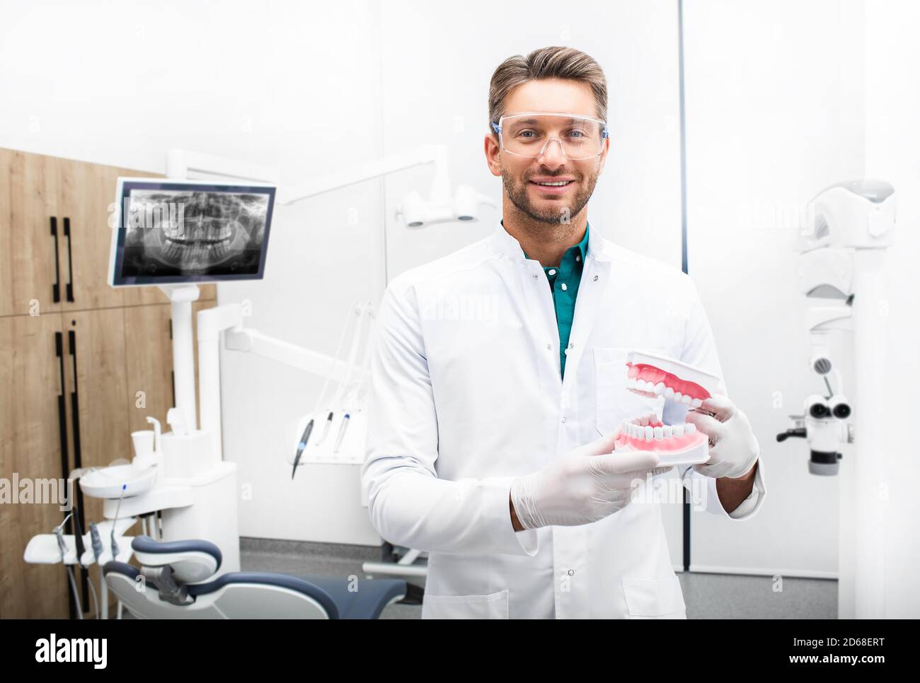 Dentista maschile sicuro in piedi nella sua clinica moderna. Igienista dentale che indossa occhiali protettivi sorridendo alla fotocamera Foto Stock
