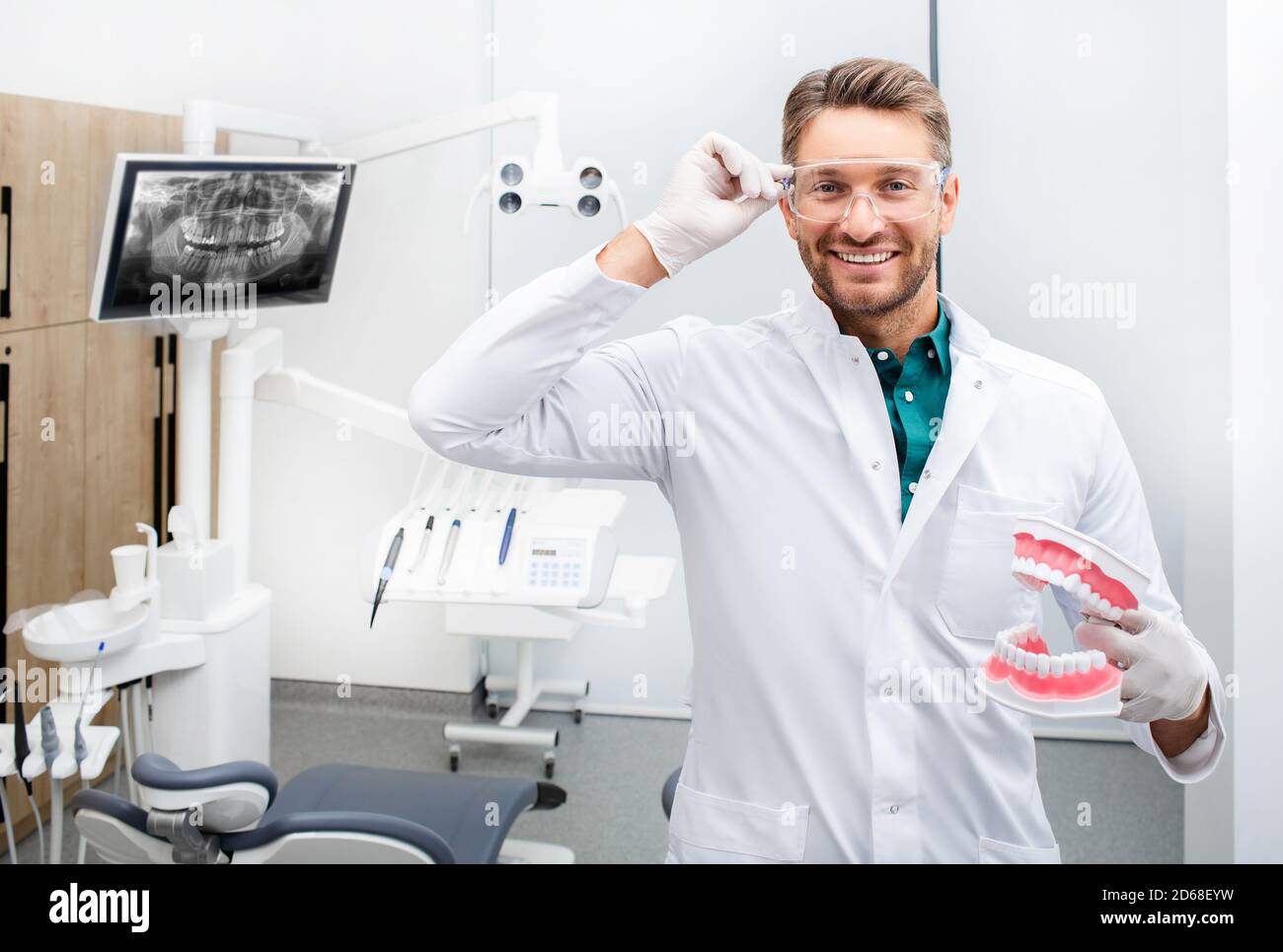 Ritratto di un bel dentista che sorride e guarda la macchina fotografica, tenendo un modello a denti. Trattamento indolore delle carie in un moderno studio dentistico Foto Stock
