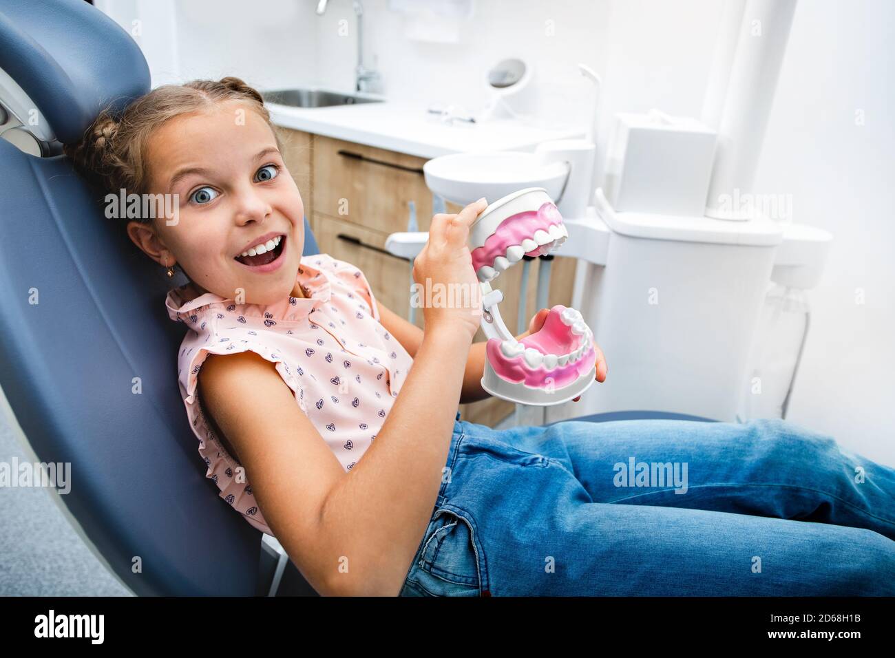 Cura dei denti dei bambini. Una bambina emozionale che gioca con un modello di mascella, si siede nella sedia del dentista. I miei denti sono sani Foto Stock