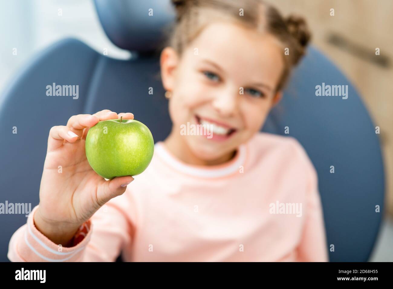 Cura dei denti dei bambini. Sorridente bambina che tiene una mela verde, si siede nella sedia del dentista. I miei denti sono sani Foto Stock