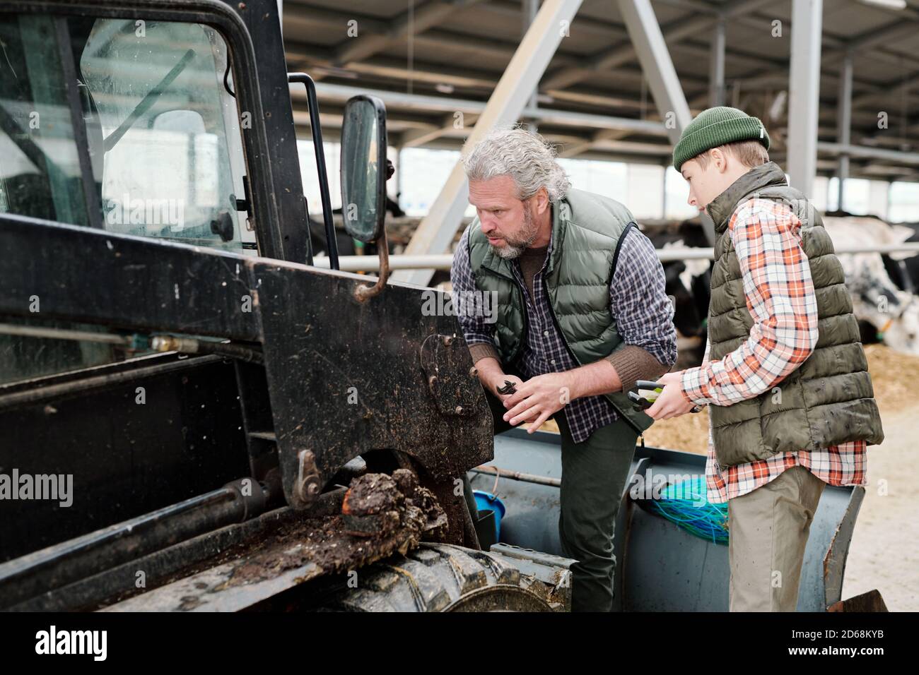 Uomo maturo concentrato con barba che analizza il motivo del guasto del trattore mentre riparandolo con il figlio alla fattoria Foto Stock