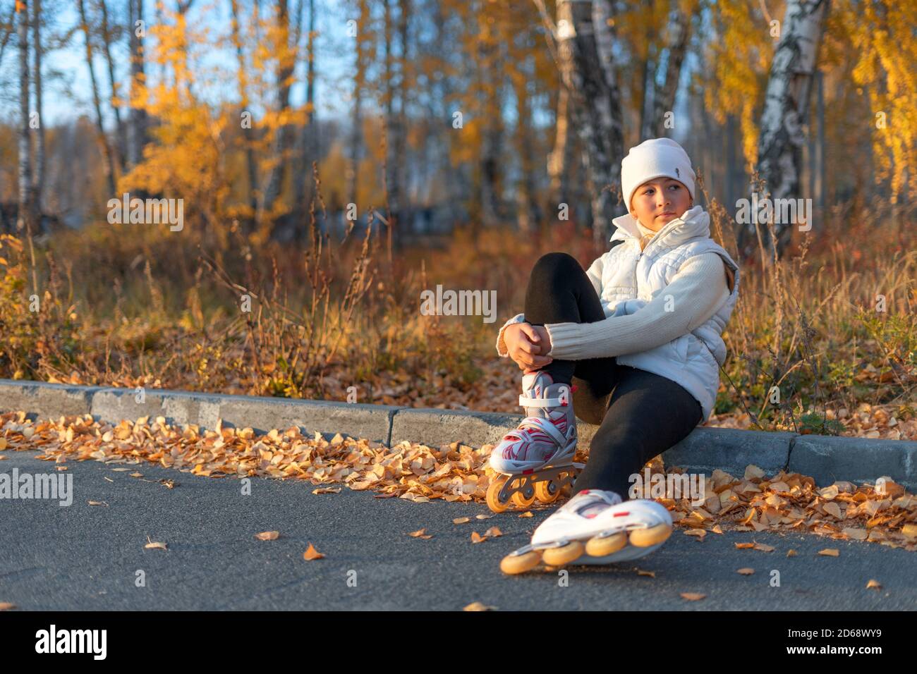 Una ragazza in pattini a rotelle è seduta su marciapiede di cemento al parco e guardare via. Un giovane skater figura sta riposando. Serata d'autunno. La ragazza indossa un cappello bianco, un maglione, una giacca e leggings neri. Spazio di copia. Foto Stock