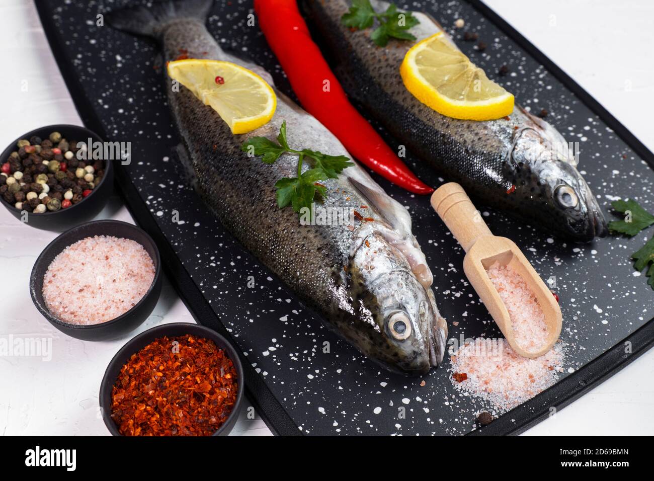 Trote iridate fresche su tavola nera con sale, pepe, limone e peperoncino ingredienti, gustosi pesci che si preparano per il pranzo Foto Stock