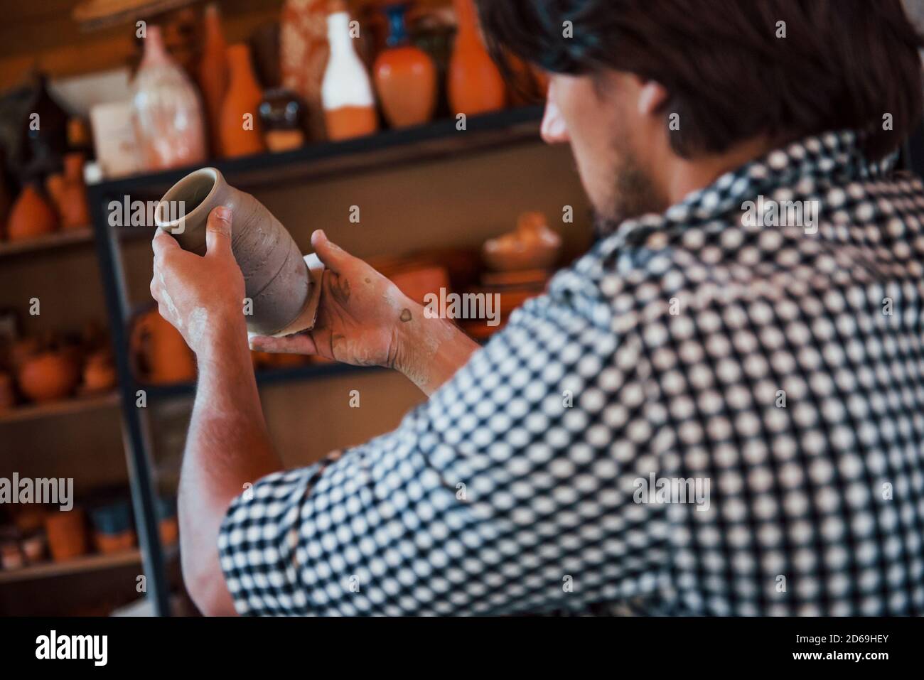 Il giovane ceramista tiene in mano una pentola fresca fatta a mano e guarda al risultato del suo lavoro Foto Stock