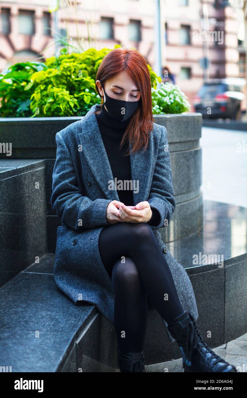 Triste donna rossa seduta in maschera protettiva in luogo pubblico e testando su un telefono. Test del coronavirus Foto Stock