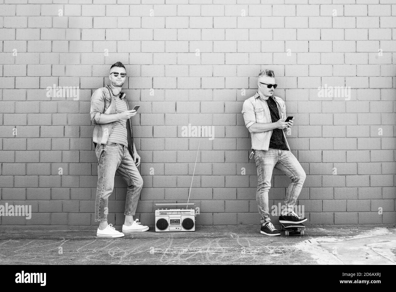 Due giovani che guardano il telefono cellulare appoggiati contro la parete di mattoni - coppia di ragazzi cool che ascoltano musica con stereo boombox vintage all'aperto in strada Foto Stock