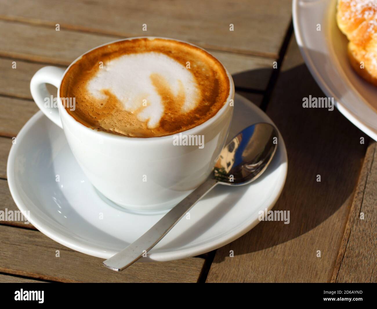 Deliziosi hot cappuccino italiano sorge in una tazza con un piattino su un tavolo di legno. Sullo sfondo di un croissant fresco su una piastra. Foto Stock