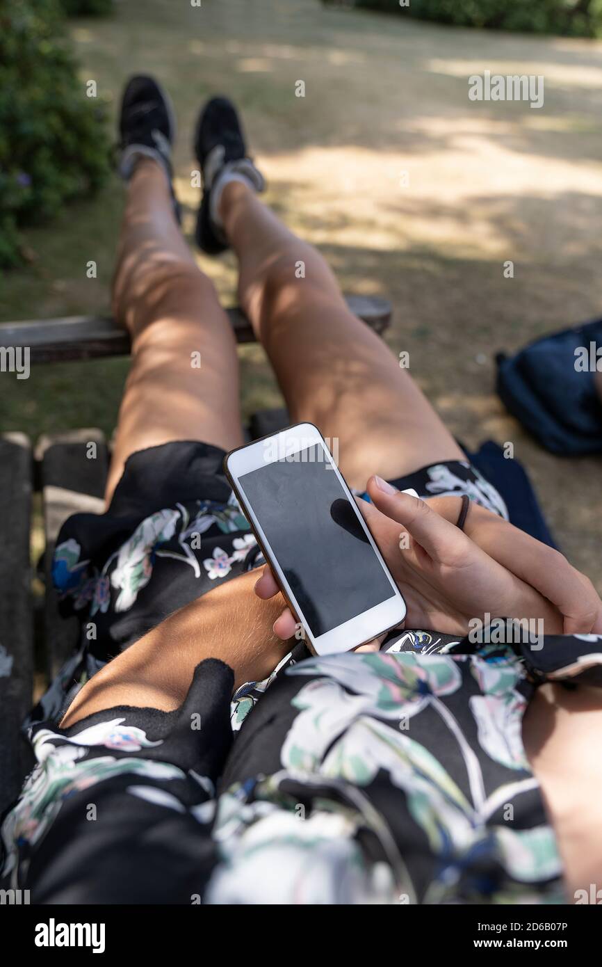 Giovane donna tiene il telefono con uno schermo nero e gioca un gioco che guarda sopra la sua spalla mentre sta mentendo giù nel parco Foto Stock