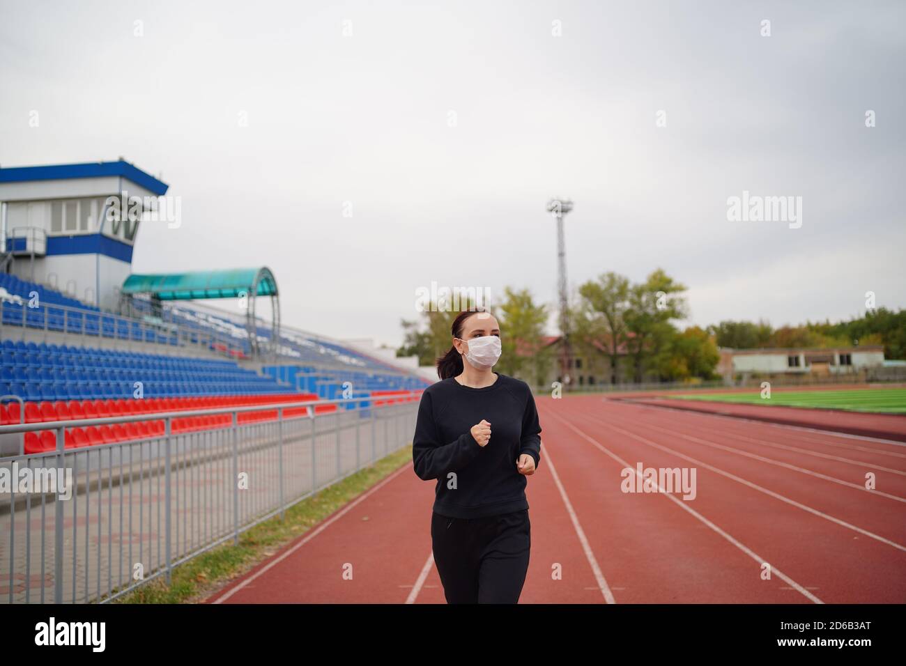 Donna attiva in maschera protettiva che corre allo stadio. Giovane fit femminile in abbigliamento sportivo e maschera protettiva per la prevenzione del coronavirus in pista rossa Foto Stock