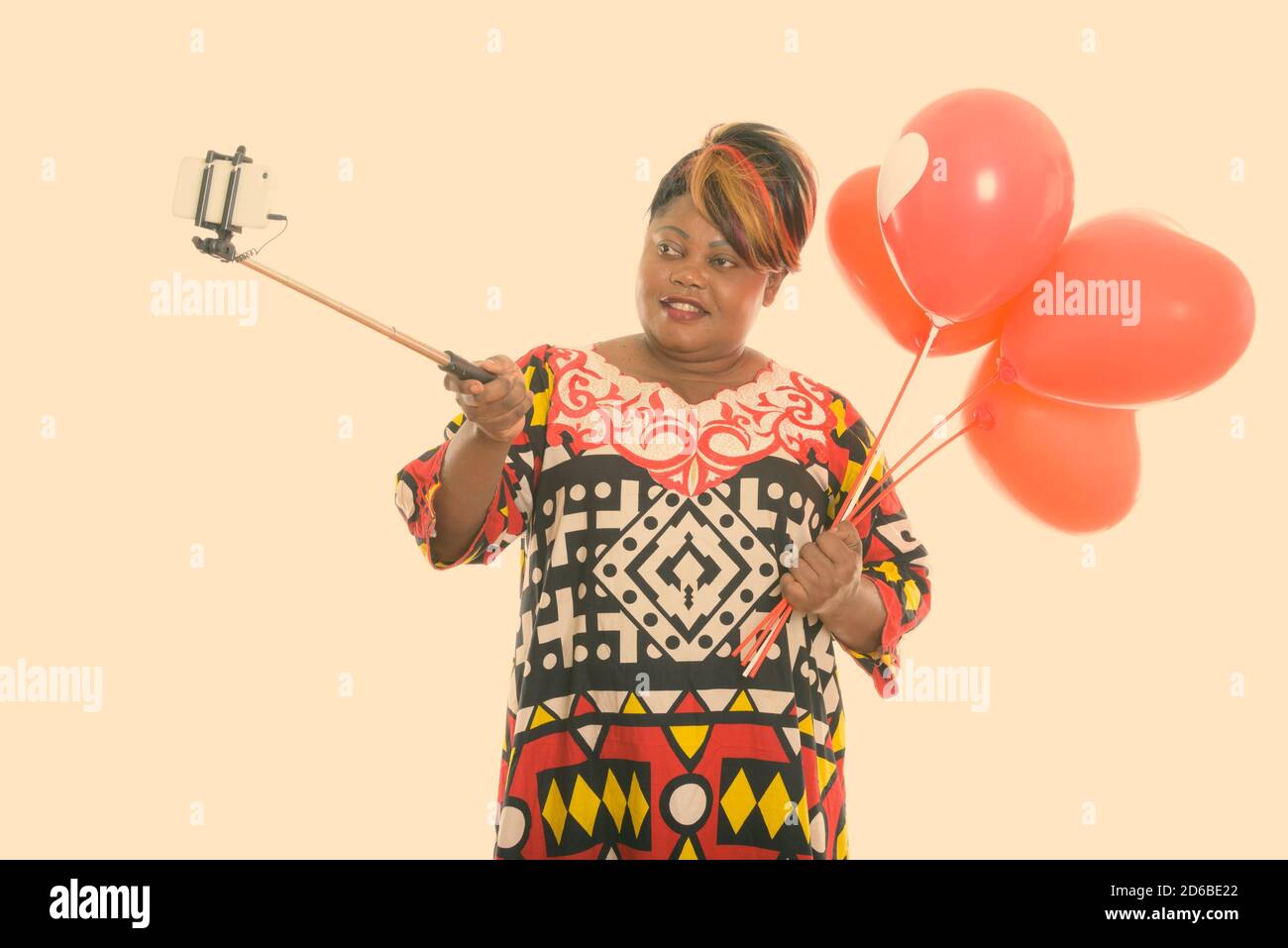 Felice grasso nero donna africana sorridente mentre tiene mazzo di palloncini rossi con il segno del cuore e scattare foto selfie con bastoncino selfie Foto Stock