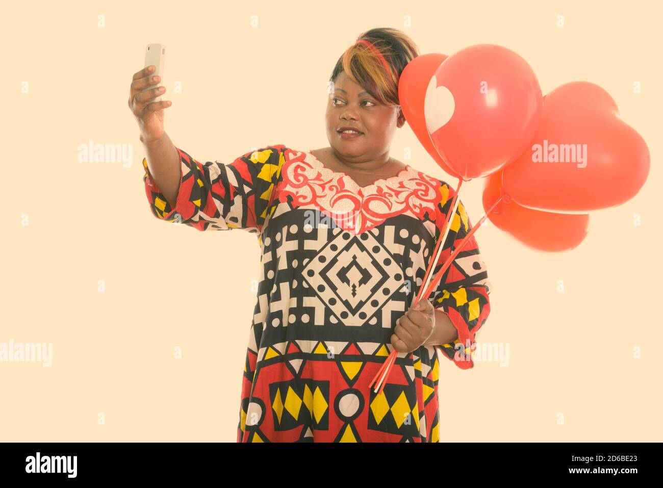 Studio di felice grasso nero donna africana sorridente mentre tenere un mazzo di palloncini rossi con il segno del cuore e prendendo immagine selfie con telefono cellulare Foto Stock