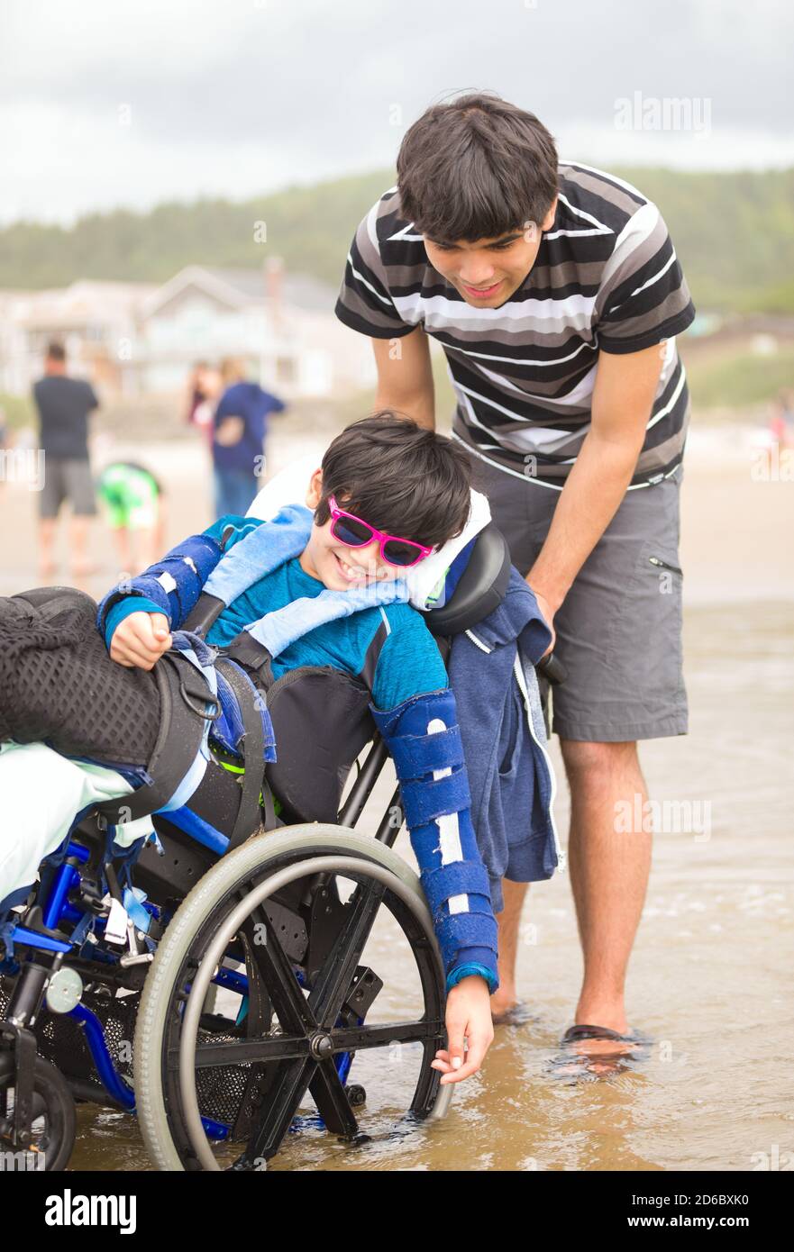 Giovane uomo caucasico asiatico biraciale che spinge il fratello minore disabile dentro sedia a rotelle sulla spiaggia Foto Stock