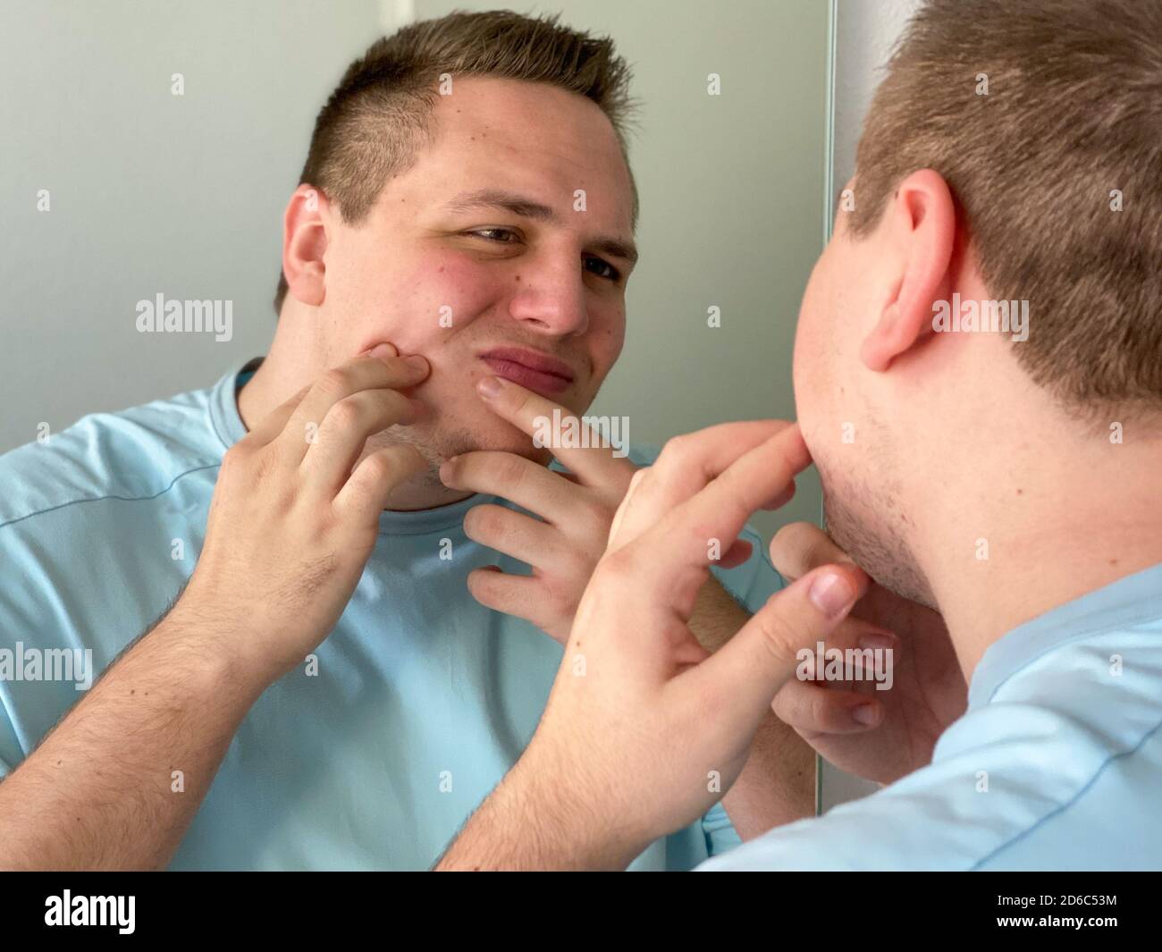 Giovane uomo che controlla il suo volto in uno specchio che cerca imperfezioni sulla sua pelle in una vista sopra la spalla e concentrarsi sulla riflessione Foto Stock