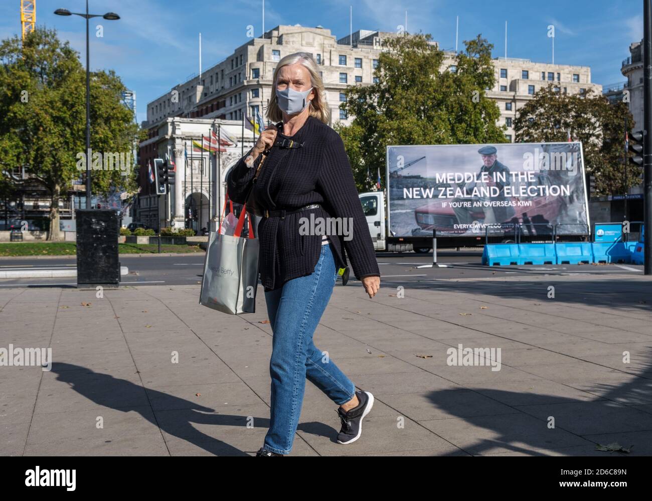 Lady con maschera facciale che tiene la borsa per lo shopping cammina a Marble Arch verso Hyde Park London. Hard Rock Hotel, bandiere, alberi e camion sullo sfondo. Foto Stock