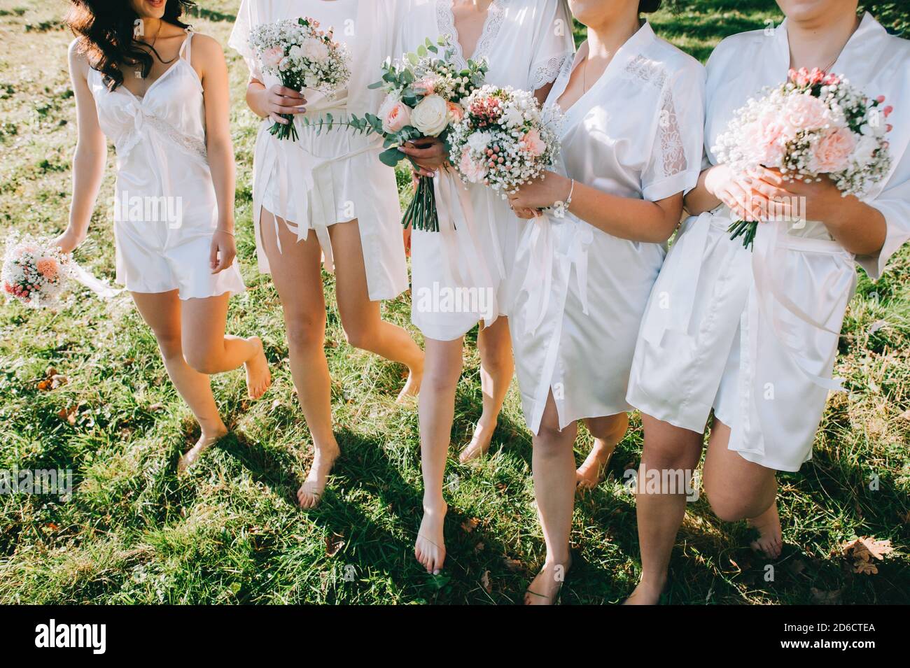 Bridesmaids senza volto e sposa in abiti satinati con bouquet in mani camminano a piedi nudi sull'erba. Foto Stock