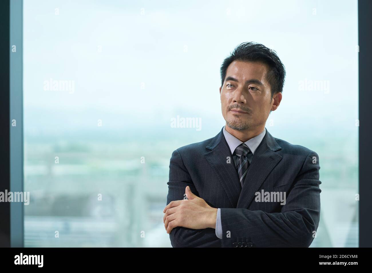 ritratto di un uomo d'affari asiatico maturo in piedi accanto alla finestra in ufficio armi incrociate Foto Stock