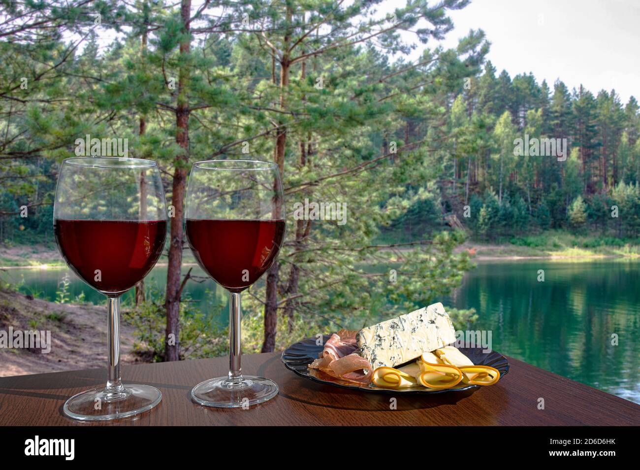 Due bicchieri di vino rosso con assortimento di salumi contro la vista di piccoli pini vicino al lago turchese Foto Stock