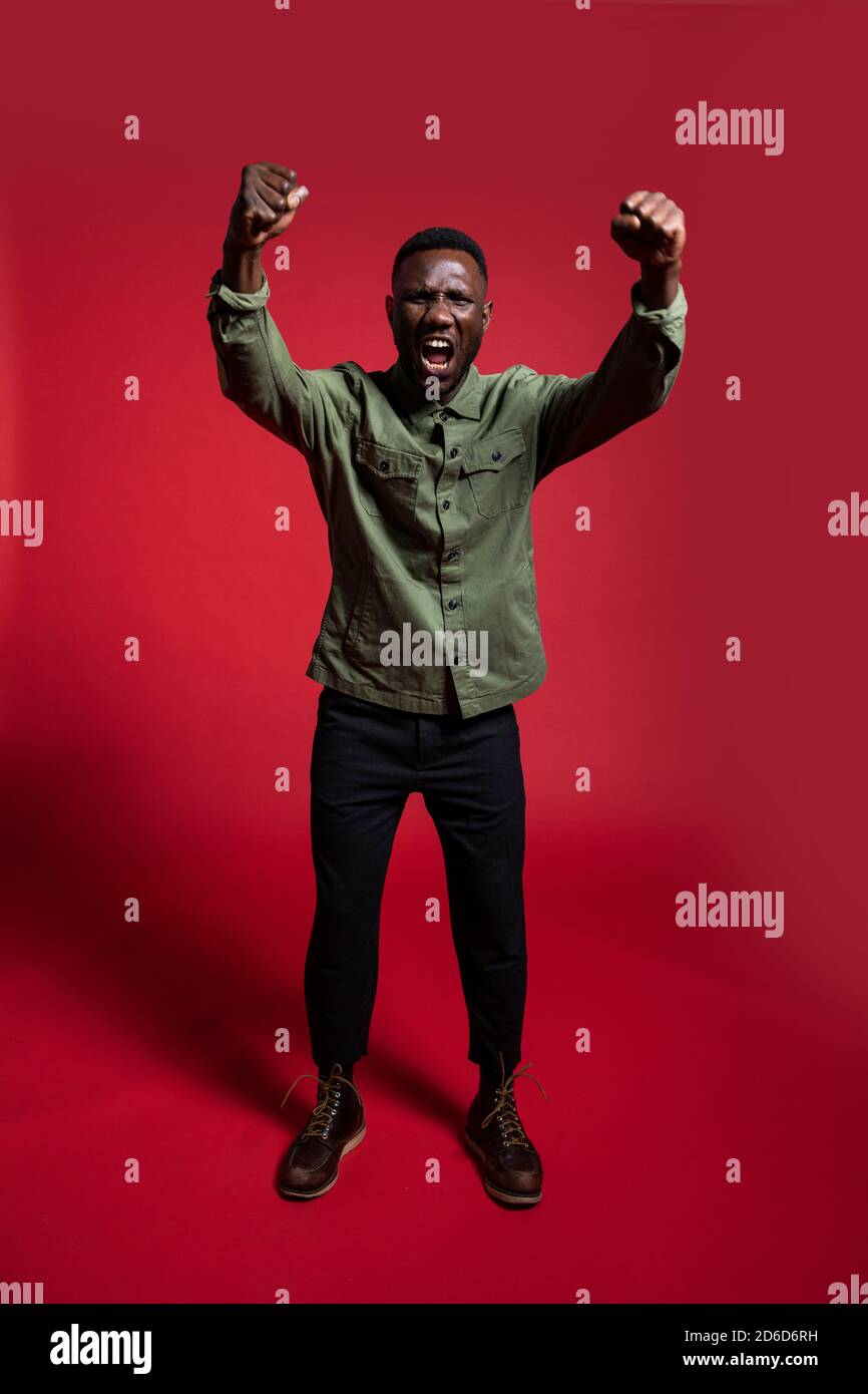 Giovane uomo nero che protesta e urla con pugni sollevati alla fotocamera per foto professionali. Isolato. Lunghezza completa. Foto Stock