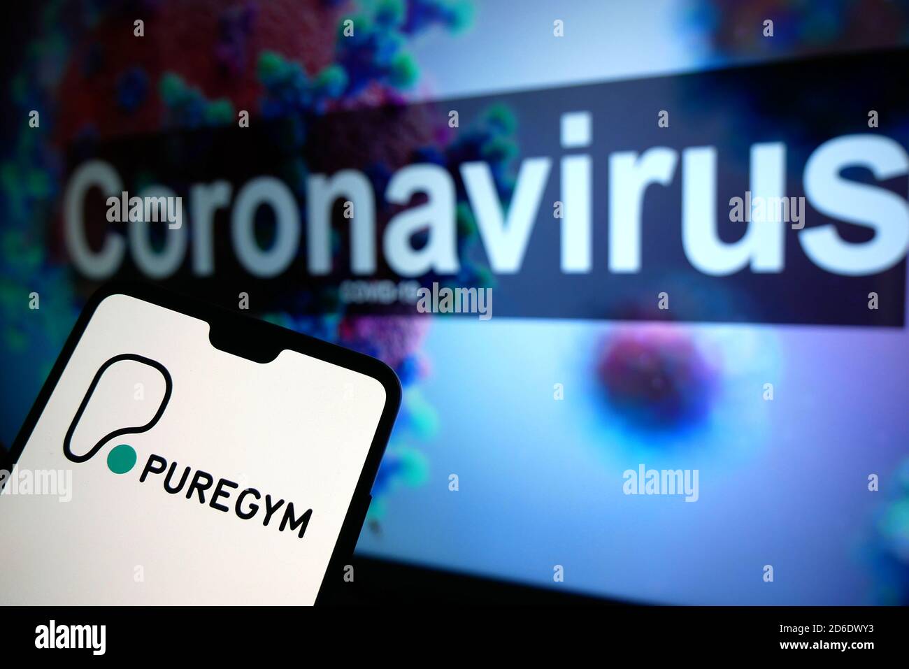 Il logo PureGym visualizzato su un telefono cellulare con un modello illustrativo del Coronavirus visualizzato su un monitor sullo sfondo. Foto Stock